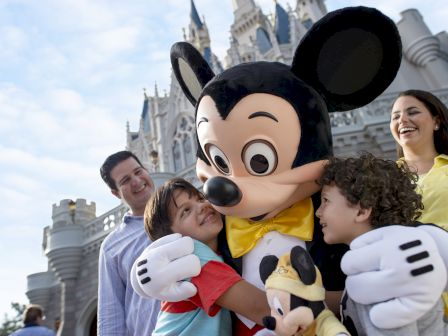 A family with two children is joyfully hugging a person dressed as Mickey Mouse in front of a castle at an amusement park.