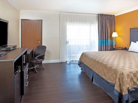 A hotel room features a king-sized bed, flat-screen TV, desk, chair, and a door. The room is well-lit with natural light from a large window.